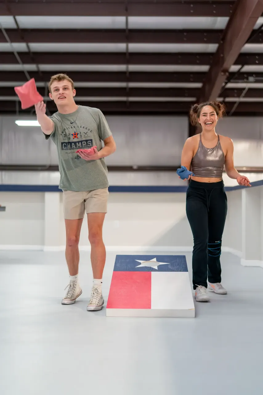 A man tosses a beanbag at a Texas-flag cornhole board while a woman laughs