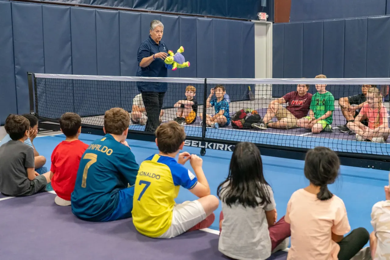 Kids sit together courtside listening to their coach at a clinic
